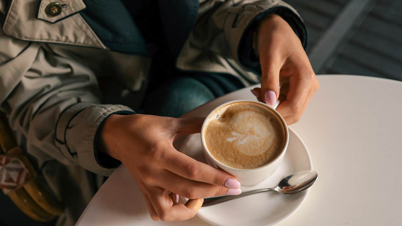 Close-up of woman's hands in trench coat gently holding a cup of cappuccino with latte art, seated at a white table